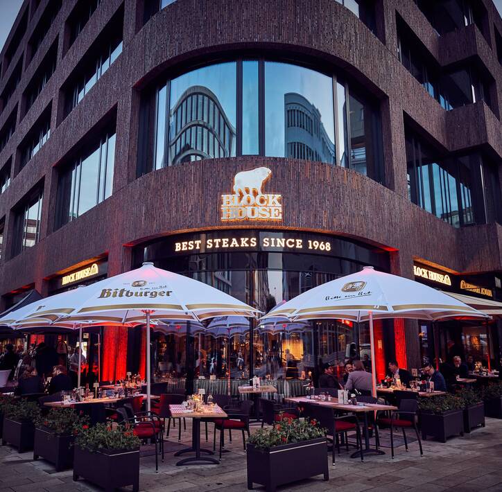 Eröffnung Deutschlandhaus Außenansicht eines modernen Block House Restaurant mit einer Terrasse davor mit Tischen und Stühlen und Schirmen